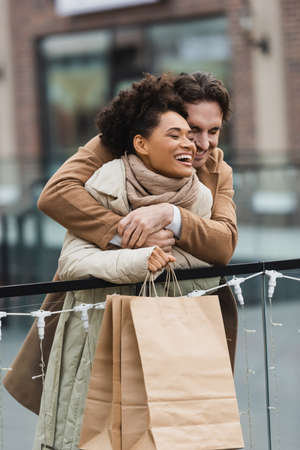 happy man hugging cheerful african american girlfriend in shopping mallの写真素材