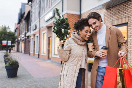 amazed interracial couple with coffee to go, christmas shopping bags and small pine near mallの写真素材