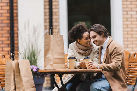 pleased african american woman holding smartphone and looking at boyfriend with paper cupの写真素材