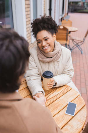 happy african american woman holding hands with blurred boyfriend near smartphone and paper cupの写真素材
