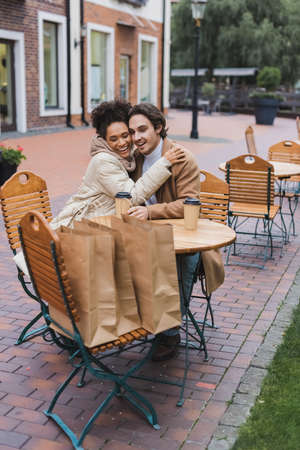 happy interracial couple embracing near paper cups and shopping bags in outdoor cafeの写真素材