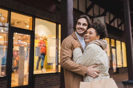 happy african american woman hugging cheerful boyfriend with shopping bagsの写真素材