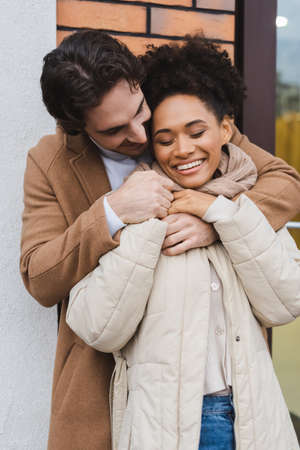 joyful man embracing cheerful african american girlfriend near building outdoorsの写真素材