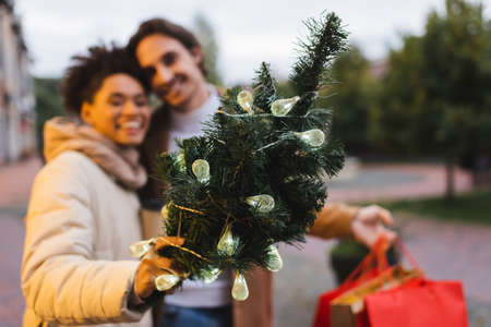 blurred african american woman holding small christmas pine with garland lights near boyfriend