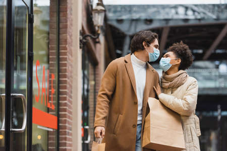 young interracial couple in medical masks looking at each other and walking with shopping bags in mallの写真素材