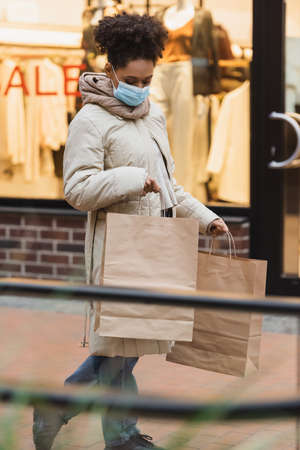 brunette african american woman in medical mask holding shopping bags in mallの写真素材