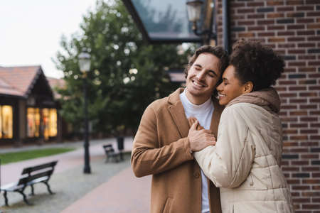 positive man embracing cheerful african american girlfriend near building outdoorsの写真素材