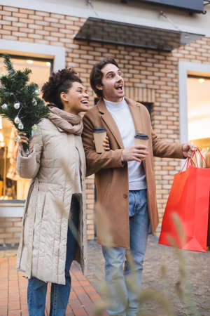 excited multiethnic couple with coffee to go, christmas shopping bag and small pine near mallの写真素材