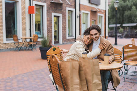 happy interracial couple hugging near paper cups and shopping bags in outdoor cafeの写真素材