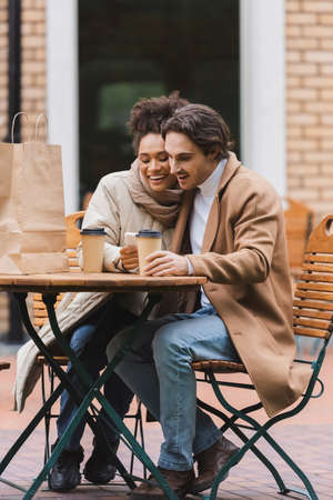 pleased african american woman holding smartphone near boyfriend with paper cup and shopping bag on tableの写真素材
