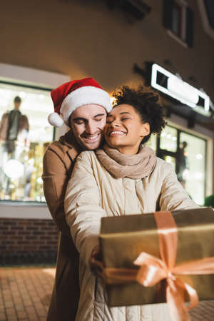 happy african american woman holding blurred wrapped gift box near boyfriend in santa hatの写真素材