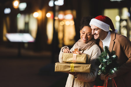 happy man in santa hat holding christmas shopping bags and small pine near african american girlfriend with gift boxesの写真素材
