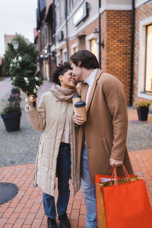 cheerful interracial couple with coffee to go, christmas shopping bags and small pine near mallの写真素材