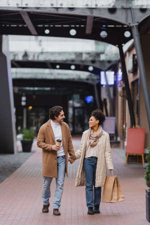 full length of african american woman holding shopping bags and walking with joyful boyfriend in coat in mallの写真素材