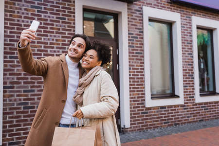 happy man taking selfie with african american girlfriend holding shopping bagsの写真素材