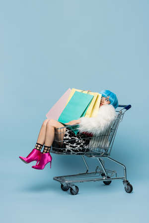 Young asian woman in pop art style sitting in shopping cart with bags on blue backgroundの写真素材