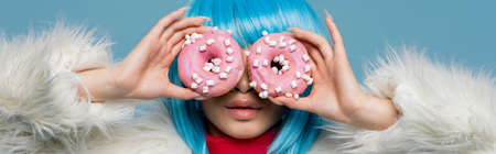 Young woman in wig and fluffy jacket holding donuts near eyes isolated on blue, bannerの写真素材
