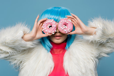 Stylish woman in bright wig holding donuts near face isolated on blueの写真素材