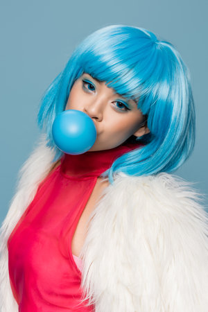 Stylish asian woman in furry jacket and wig blowing bubble gum isolated on blueの写真素材