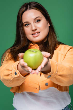 Pretty woman with overweight holding blurred apple and looking at camera isolated on greenの写真素材