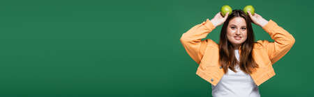 Cheerful body positive woman holding apples near head isolated on green, bannerの写真素材