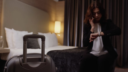 suitcase near blurred businesswoman talking on smartphone and looking at wristwatch in hotel roomの写真素材