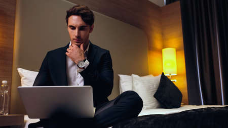 young pensive businessman sitting on bed and using laptop in hotel roomの写真素材