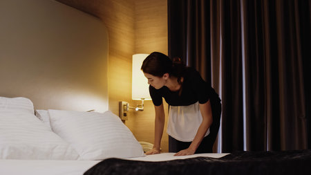 young maid in uniform making bed in hotel roomの写真素材