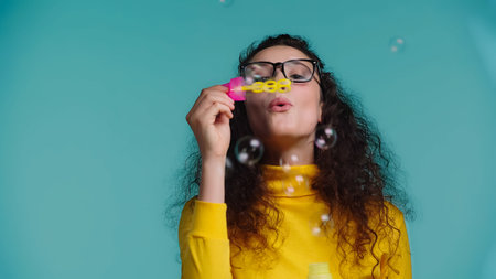 young woman in glasses blowing soap bubbles isolated on blueの写真素材