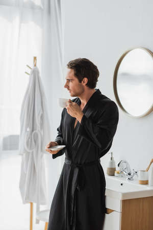 Side view of young man in silk robe holding cup and saucer in bathroom in morningの写真素材