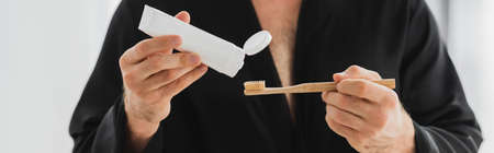 Cropped view of man in robe holding toothpaste and toothbrush at home, banner