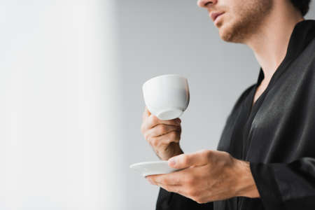Cropped view of cup and saucer in hands of young man in silk robe at home