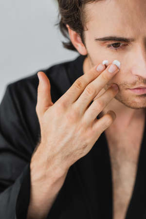 Cropped view of young man in black robe applying cosmetic cream on face isolated on greyの写真素材