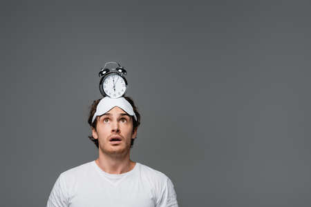 Shocked man in sleep mask looking at alarm clock on head isolated on grey