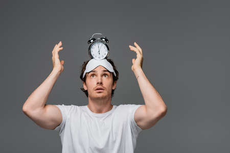 Young man in sleep mask and white t-shirt holding alarm clock on head isolated on greyの写真素材