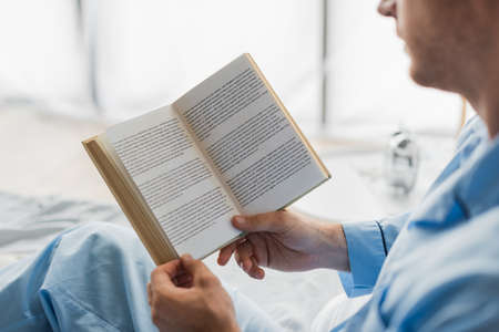 Cropped view of book in hands of blurred man in pajama on bedの写真素材