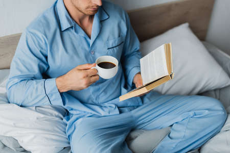 Cropped view of man in pajama holding coffee cup while reading book on bed