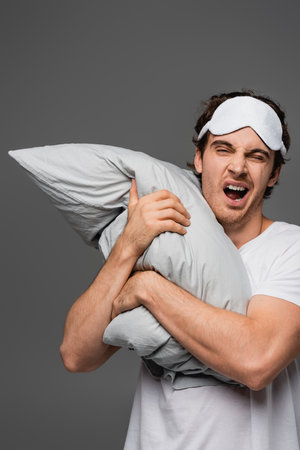 Man in sleep mask yawning while holding pillow isolated on greyの写真素材