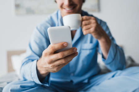 Cropped view of smartphone in hand of blurred man with cup in bedroomの写真素材