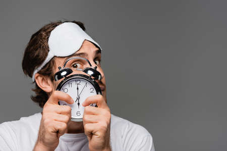 Young man in sleep mask covering face with alarm clock isolated on greyの写真素材