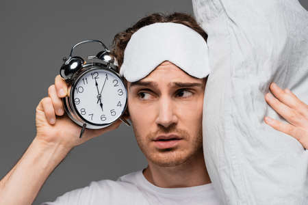 Young man in sleep mask holding pillow and alarm clock isolated on greyの写真素材