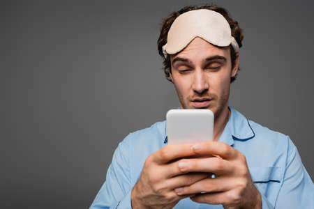 Sleepy man in pajama using smartphone isolated on greyの写真素材