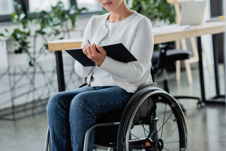cropped view of businesswoman in wheelchair writing in notebook in officeの写真素材
