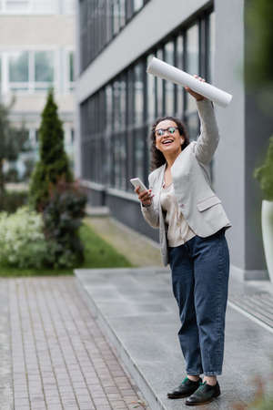cheerful african american businesswoman with mobile phone looking away and waving rolled papers near blurred buildingの写真素材