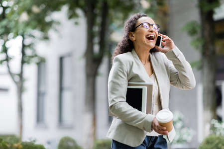 laughing african american businesswoman with takeaway drink, digital tablet and documents talking on...