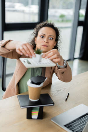 african american businesswoman with funny grimace making pyramid from flowerpot, stationery and paper cup on deskの写真素材