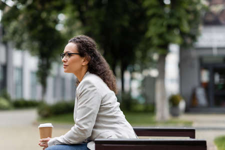 side view of african american businesswoman in eyeglasses sitting with coffee to go outdoorsの写真素材