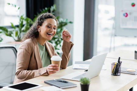 african american businesswoman with coffee to go smiling during video call on laptop in officeの写真素材