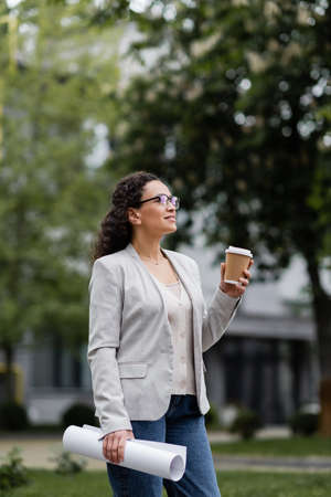 african american businesswoman with rolled documents and takeaway drink looking away outdoorsの写真素材