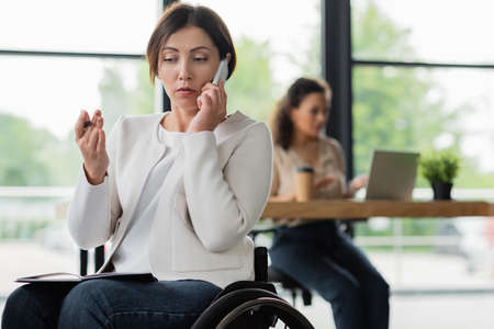 businesswoman in wheelchair talking on mobile phone near african american woman working on blurred backgroundの写真素材